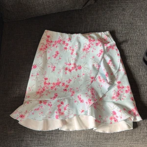 Blommig omlottkjol från NLY One XS - Supersöt kort omlottkjol från NLY One i ljusblått med rosa blommor. Kjolen har volangkant och en romantisk vibe, perfekt för vår och sommar. Sitter skönt i midjan och är lätt att matcha med en enkel topp.