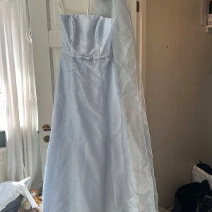 Ljusblå långklänning med pärldetaljer samt sjalett - Elegant vit långklänning med axelbandslös design och subtila pärldetaljer på överdelen. Klänningen har en liten rosett i midjan och ett skirt, svepande tyg som ger en drömmig känsla. Perfekt för speciella tillfällen när du vill känna dig extra fin.