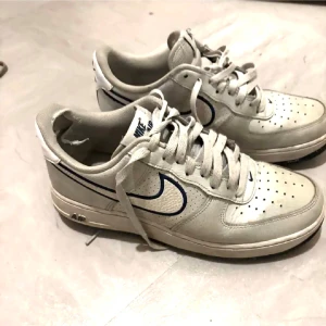 Nike Air Force 1 vita sneakers - Klassiska Nike Air Force 1 sneakers i vitt skinn med blå detaljer och perforerad tå. Lågt skaft, snörning och ikonisk swoosh-logga på sidan. Perfekta för en clean och sportig look.