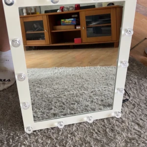Sminkspegel med lampor  - Säljer en vit sminkspegel med lampor runt. Spegeln har rektangulär form och kan både stå på bordet och hängas på väggen. längden är 45cm och bredden 35cm