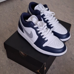Nike Air Jordan 1 Low vit/marinblå - Snygga Nike Air Jordan 1 Low sneakers i vitt och marinblått med grå swoosh. Klassisk siluett med perforerad tå, vita skosnören och låg profil. Ovandel i skinn och syntet, perfekt för dig som gillar streetstyle och vill sticka ut med en ikonisk modell.