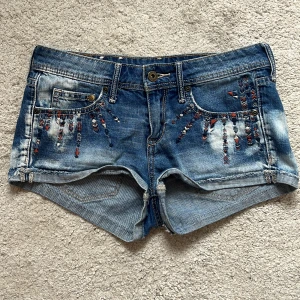 Lågmidjade jeansshorts - Snygga blå jeansshorts från H&M med broderade detaljer i rött, vitt och blått framtill och på ena bakfickan. Klassisk femficksmodell med låg midja och slitna kanter. Perfekta för sommaren och ger en cool, avslappnad vibe.