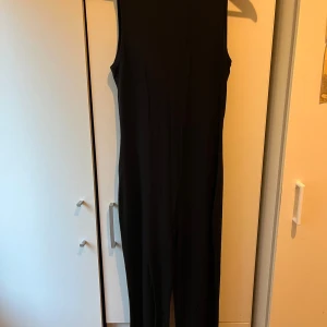 Svart ärmlös jumpsuit - Säljer en svart, ärmlös jumpsuit med smal passform och rund halsringning. Perfekt för dig som gillar en clean och stilren look. Materialet känns mjukt och följsamt mot kroppen. Enkel att styla med sneakers eller klackar.