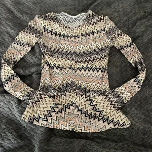 Zigzag-mönstrad topp från KappAhl - Snygg långärmad topp från KappAhl med färgglatt zigzag-mönster i svart, beige, gul och rosa. Figurnära passform och knappar framtill. Perfekt för dig som gillar unika mönster och vill sticka ut lite extra.