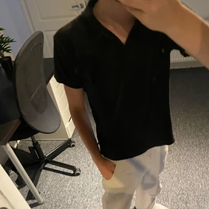 Svart frottéskjorta - Svart kortärmad skjorta från Kappahl i mjuk frotté. Skjortan har klassisk krage och rak passform, perfekt för en chill och avslappnad stil. Enkel att matcha med både jeans och shorts för en clean look. Modellen är 162 och är kort men ändå hyffsad i gemförelse