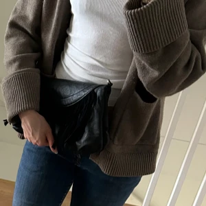 Svart väska/clutch med fransar  - Säljer en svart väska i skinn med snygga fransdetaljer på framsidan. Väskan har avtagbar axelrem och kan bäras både på axeln och som clutch. Perfekt storlek för det viktigaste och ger en cool touch till din outfit.