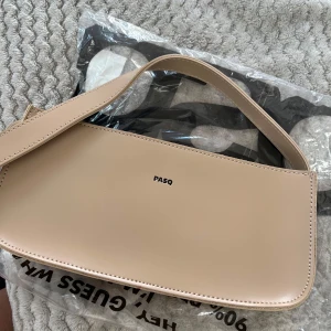 Beige axelväska från PASQ - Snygg beige axelväska från PASQ i stilren design. Väskan har ett minimalistiskt utseende med slät yta, bred axelrem och dragkedja upptill. Perfekt storlek som passar till många olika outfits.