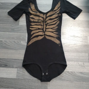 Svart body med guldmönster, skelett från Monki - Cool svart body från Monki i storlek XS med korta ärmar och ett unikt guldmönster framtill som påminner om ett skelett eller ribbor. Bodyn har rundad urringning och knäppning nedtill. Perfekt för dig som vill sticka ut med en edgy vibe. Lite tvättpåverkat men fortfarande väldigt fin! 