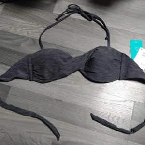 Ny sexig, mörkgrå bikiniöverdel från H&M - Snygg mörkgrå bikiniöverdel från H&M med twistad front.. Bikinin har vadderade kupor och knytning i nacken och ryggen. Perfekt för strandhäng och poolparty. Materialet är mjukt och stretchigt för bästa passform.