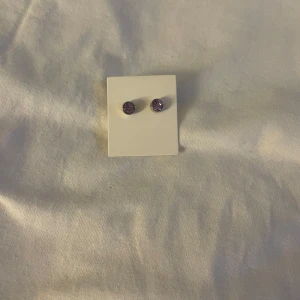 Lila studs örhängen med sten - Säljer ett par små, runda studs örhängen med lila glittrande sten. Klassisk design med silverfärgad baksida och enkel push-back låsning. Perfekta för dig som gillar en subtil men färgglad detalj i din look.