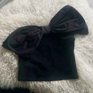 Svart tubtopp med stor rosett - Supersnygg svart tubtopp med en riktigt stor rosett över bysten. Toppen är axelbandslös och har en tight passform som framhäver figuren. Perfekt för dig som vill sticka ut och skapa en cool look.