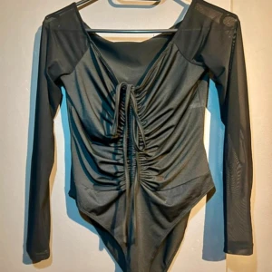 Svart mesh body med snörning - Snygg svart body med långa meshärmar och trendig snörning framtill som ger en cool rynkad effekt. Djup v-ringning och åtsittande passform. Perfekt att styla med jeans eller kjol för en edgy look.