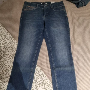 MAC Angela blå jeans med glitterdetalj - Snygga blå jeans från MAC, modell Angela, med raka ben och glittriga detaljer på fickorna. Jeansen har klassisk femficksdesign och är tillverkade i mjukt bomullstyg med en touch av stretch för extra komfort.