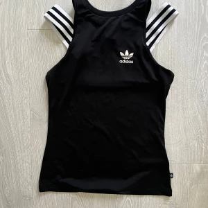 Svart linne från Adidas med ränder - Snyggt svart linne från Adidas med klassiska vita ränder på axelbanden och Adidas-logga framtill. Modellen är ärmlös och har en sportig vibe, perfekt för dig som gillar streetstyle eller träning. Materialet känns mjukt och följsamt.