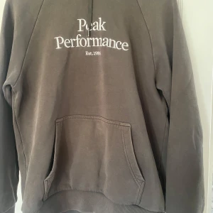 Grön hoodie från Peak Performance - Säljer en grön hoodie från Peak Performance med broderad logga på bröstet. Tröjan har huva med snörning, känguruficka och långa ärmar. Perfekt för chill dagar och enkel att matcha med jeans eller joggers.  ( på bilderna ser den brun ut men den är mörk grön)