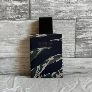 Replay Signature Eau de Toilette - Parfymflaska från Replay Signature med coolt kamouflagemönster i grönt, svart, beige och grått. Flaskan är rektangulär med räfflade sidor och har en stor svart fyrkantig kork med Replay-loggan. Tillverkad i Italien, 100 ml Eau de Toilette.