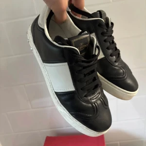 Valentino sneakers i svart och vitt - Säljer ett par svarta och vita Valentino sneakers med klassisk design och silverfärgade nitar på hälen. Skorna har snörning och är tillverkade i skinn. Perfekta för dig som gillar stilrena och exklusiva sneakers. St 38 men passar 39/40