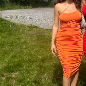 Orange tight midiklänning med drapering - Säljer en orange, figurnära midiklänning med draperad effekt och asymmetrisk axelband. Klänningen är ärmlös och har en snygg, kroppsnära passform som framhäver siluetten. Perfekt för dig som vill sticka ut med en färgstark look.