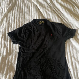 Svart t-shirt från Polo Ralph Lauren - Svart Ralph Lauren T-shirt - Storlek M Snygg och stilren svart T-shirt från Polo Ralph Lauren med klassisk röd logga på bröstet. Passar perfekt både till vardags och festligare tillfällen. Mjuk bomullskvalitet och bekväm passform. Storlek: M Skick: Bra använt skick – inga hål eller fläckar.