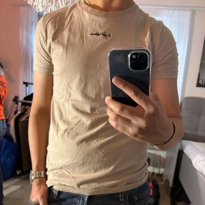 Beige t-shirt från Hollister - Snygg beige t-shirt från Hollister Must-Have Collection med svart broderad logga på bröstet. Klassisk rund halsringning och korta ärmar. Tillverkad i 100% bomull för en skön och lätt känsla. Perfekt till jeans eller shorts för en clean look.