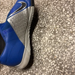 Blå Nike fotbollsskor med silverdetaljer - Snygga blå Nike fotbollsskor med silverfärgade detaljer och svart/vit swoosh. Skorna har en strumpliknande svart öppning och är tillverkade i syntetmaterial för bra passform och känsla på planen. Perfekta för dig som vill sticka ut på fotbollen.