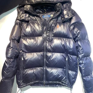 Mörkblå pufferjacka Polo Ralph Lauren - Snygg mörkblå pufferjacka från Polo Ralph Lauren med huva och flera dragkedjefickor. Jackan har klassisk broderad logga på bröstet och är vadderad för extra värme. Perfekt för kalla dagar och har en clean, sportig look.