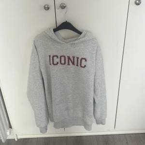 Grå hoodie ICONIC från Gina Tricot - Grå hoodie från Gina Tricot med vinröd text 'ICONIC' broderad på bröstet. Mjuk insida, huva och långa ärmar. Perfekt för chill dagar och enkel att matcha med jeans eller leggings.