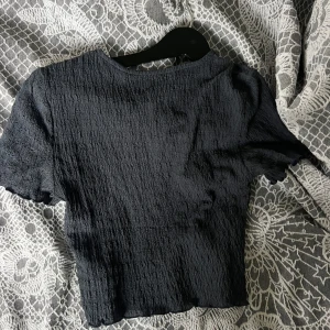 Svart croppad topp från Vero Moda - En svart croppad topp från Vero Moda med korta ärmar och en lätt skrynklig struktur. Toppen har en enkel, rund halsringning och är tillverkad i ett mjukt, stretchigt material som sitter snyggt på kroppen. Perfekt för att lyfta vilken outfit som helst.