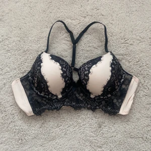 Svart spets-bh från Hunkemöller - Supersnygg bh från Hunkemöller i svart spets med ljusrosa underton. Dekorerad med en liten rosett framtill och fina spetsdetaljer över kuporna. Axelbanden är justerbara och modellen ger ett riktigt elegant intryck.