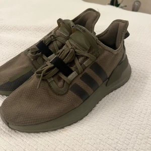 Olivgröna Adidas sneakers med snörning - Snygga olivgröna Adidas sneakers med klassiska tre ränder på sidan. Ovandelen är i mesh-liknande syntetmaterial och skorna har snörning samt platt sula. Perfekta för dig som gillar en stilren och sportig look. (Helt oanvända)