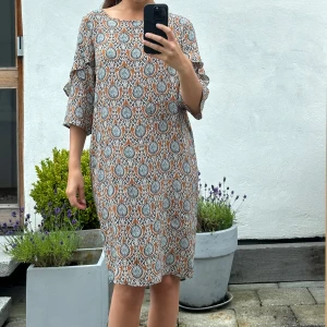 Mönstrad kortklänning med volangärm - Säljer en kortklänning med unikt mönster i brunt, svart, vitt och blått. Klänningen har trekvartsärm med volangdetalj och en rak, loose passform. Materialet känns lätt och luftigt, perfekt för varmare dagar. Rund halsringning och snyggt fall.