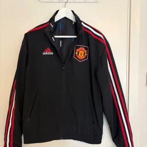 Manchester United vindjacka Adidas - Svart Manchester United vindjacka från Adidas med röda och vita ränder. Nypris 1300kr, skick 9,5/10. Storlek S.