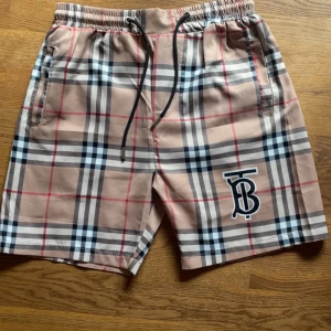 Rutig Burberry shorts med snörning - Snygga shorts från Burberry med klassiskt rutig mönster i beige, svart, vit och röd. De har elastisk midja med snörning och sidofickor samt Burberrys TB-logga framtill. Perfekta för sommaren och ger en lyxig vibe till din outfit. Hör gärna av er vid frågor 