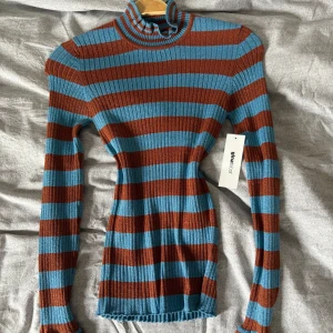 Randig tröja från Gina Tricot - Supersnygg ribbad polotröja från Gina Tricot i blått och brunt glittrande tyg. Tröjan har lång ärm, smal passform och coola volangdetaljer i kragen. Perfekt för dig som gillar retrovibbar och vill sticka ut med färg. Stretchigt material som sitter tajt men bekvämt, materialet sticker inte. Helt oanvänd med lappar kvar. 