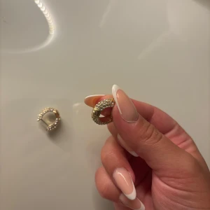 Guldiga örhängen från Pilgram - Säljer ett par chunky guldiga örhängen från pilgram, täckta med gnistrande vita stenar. De är runda och har en lyxig känsla med glittrig yta som verkligen drar blickarna till sig. Perfekta för dig som vill ha något extra till din look.