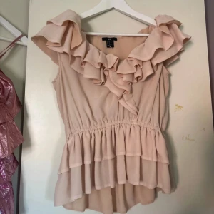 Volang topp - Supersöt beige blus från H&M med stora volanger runt halsen och dubbla volanglager nertill. Ärmlös modell med markerad midja och flowig passform. Perfekt för dig som gillar romantiska vibes och vill sticka ut med detaljer.