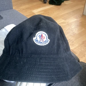 Svart bucket hat från Moncler - Snygg svart bucket hat från Moncler med klassisk broderad logga framtill i vitt, blått och rött. Hatten är tillverkad i bomull och har en mjuk, rundad form med bred brätte. Perfekt accessoar för en clean och trendig look.