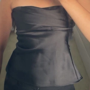 Zara svart satin bandeau tubtopp - Snygg svart bandeau tubtopp i glansigt satinmaterial. Toppen har en stilren, axelbandslös design med draperad detalj upptill som ger en lyxig känsla. Perfekt att styla med jeans eller kjol för en trendig look.