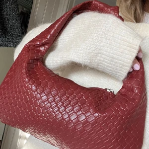 Vinröd axelväska  - Säljer en vinröd axelväska i flätat skinn med mjuk, rundad form. Väskan har en bred axelrem med knutdetalj och stängs med dragkedja upptill. Perfekt storlek för det viktigaste och ger en cool touch till din outfit.