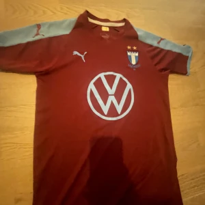 Vinröd Malmö FF fotbollströja Puma - Snygg vinröd Malmö FF fotbollströja från Puma med ljusblå detaljer på ärmarna och stora VW-loggan på bröstet. Klassisk klubbsköld med två stjärnor och Puma-logga på bröst och ärmar. Tillverkad i lätt och ventilerande material.