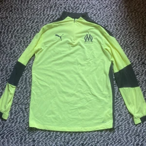 Marseille tracksuit - Snygg långärmad fotbollströja från Olympique de Marseille i neongult och svart. Tröjan har halv dragkedja, klubbmärke och Puma-logga på bröstet. Tillverkad i lätt och ventilerande funktionsmaterial, perfekt för träning eller match.
