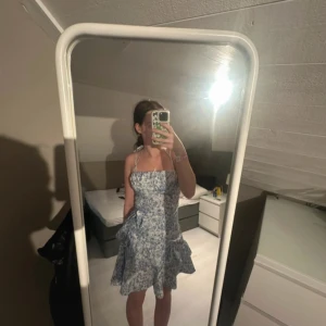 Blåvit blommig kort sommarklänning - Supersöt blåvit kort klänning med blommigt mönster och volangkant. Klänningen har smala axelband som knyts i nacken och en lätt utsvängd kjol. Perfekt för varma dagar och ger en fräsch och ungdomlig look.