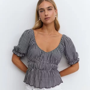 Svartvit rutig blus med puffärm - Supersöt blus i svartvitt rutigt mönster med djup urringning och puffiga korta ärmar. Smockad midja ger en snygg siluett och volangkant nertill. Perfekt för dig som gillar trendiga detaljer och klassisk stil. Endast testad.