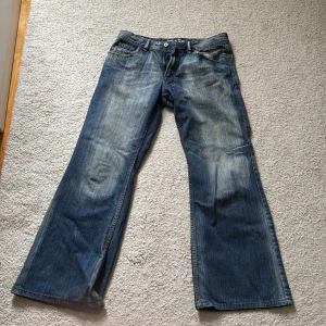 Blå bootcut jeans från Seven7 - Snygga blå jeans från Seven7 med bootcut passform och klassisk femficksdesign. Jeansen har slitningar och ljusare partier framtill och bak, samt broderade detaljer på bakfickorna. Perfekta för dig som gillar en avslappnad och trendig look.