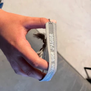 Grått Christian Dior armband med tofsar - Snyggt vävt armband från Christian Dior i ljusgrått med vit broderad logga. Armbandet har justerbar knytning och dekorativa tofsar i ändarna. Perfekt accessoar för att lyfta din outfit med en exklusiv touch.