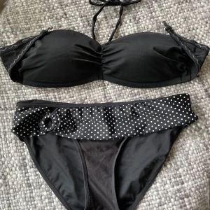 Svart bikini med prickiga detaljer - Svart bikini med bandeau-topp som har knytband i nacken och lätt rynk framtill. Bikinitrosan har en svart bas och en bred volangkant upptill i svart med vita prickar samt en liten rosett på sidan. 