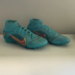 Nike blå fotbollsskor elite - Säljer ett par sjukt snygga blå Nike fotbollsskor med högt skaft och snörning. Skorna har en cool design med orange och gul detalj på swoosh-loggan och mönstrad sula. Ovandelen är i syntetmaterial och skon har en tight passform som ger bra känsla på planen.