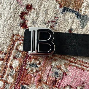 Burberry vändbart skärp med B-spänne - Snyggt vändbart Burberry-bälte i skinn med ikoniskt rutigt mönster på ena sidan i beige, brun och röd samt helsvart på andra sidan. Stort silvrigt B-spänne framtill. Perfekt accessoar för att lyfta din outfit med klassisk Burberry-stil.