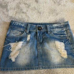 Blå jeanskjol med slitningar och pärlor - Snygg blå jeanskjol i kort modell med slitna detaljer och pärlor på både fram- och baksidan. Kjolen har klassiska fickor, bälteshällor och en cool, ruffig look. Perfekt för dig som gillar streetstyle och vill sticka ut lite extra.