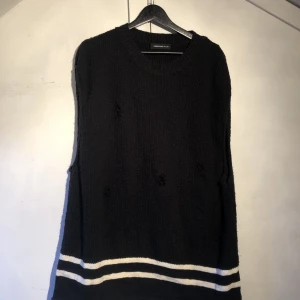 Svart ribbstickad oversize vest/linne från undercover - Svart stickad vest/linne från Undercover med vita ränder längst ner. har en relaxed fit, rund halsringning och slitna detaljer framtill. Materialet är 78% bomull och 22% akryl, vilket gör den både mjuk och lite chunky. Perfekt för dig som gillar streetwear med edge.